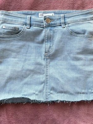 Ljusblå jeanskjol med rå kant - Trendig ljusblå jeanskjol från Jane Skirt i klassisk femficksmodell. Kjolen har en rå, fransig kant nertill och bälteshällor i midjan. Perfekt för dig som gillar en avslappnad och cool stil.