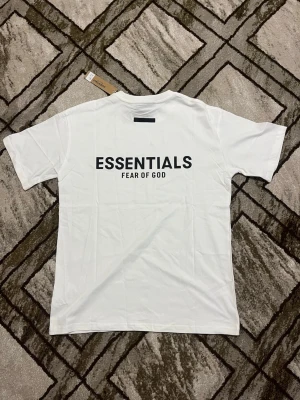 Vit Essentials Fear of God t-shirt - Säljer en vit t-shirt från Fear of God Essentials med svart tryck på ryggen. Klassisk rund hals och korta ärmar. Tillverkad i mjuk bomull för en clean och stilren look. Perfekt för dig som gillar streetwear och minimalistisk design.