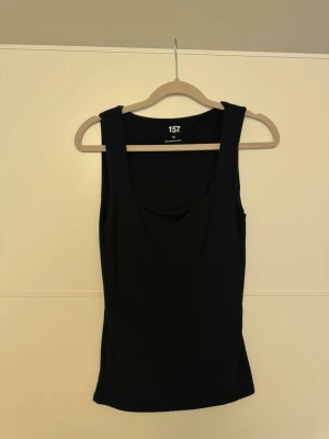 Svart tanktop från 157 - Snygg svart tajt tanktop från 157 med breda axelband och rundad halsringning. Tillverkad i mjuk och stretchig viskosblandning (95% viskos, 5% elastan) som sitter skönt på kroppen. Perfekt att styla lager på lager eller bära som den är.☀️