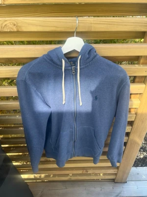Blå hoodie från Polo Ralph Lauren - Mysig blå hoodie från Polo Ralph Lauren med dragkedja framtill, vit dragsko i huvan och klassisk logga broderad på bröstet. Hoodien har känguruficka och är tillverkad i mjukt bomullsmaterial. Perfekt för en avslappnad stil.