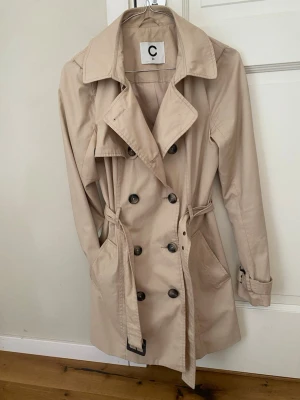 Beige trenchcoat med bälte - Stilren beige trenchcoat med dubbelknäppning, klassisk krage och bälte i midjan. Jackan har två sidfickor och justerbara ärmslut. Perfekt för vår och höst när du vill ha en tidlös look.