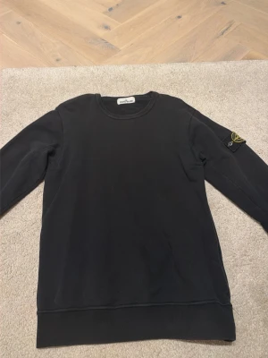 ÄKTA Svart sweatshirt från Stone Island - Svart sweatshirt från Stone Island med klassisk rund halsringning och ikonisk logotyp-patch på vänster ärm. Tröjan har en enkel och stilren design, perfekt för en avslappnad streetwear-look. Tillverkad i mjukt bomullsmaterial och har långa ärmar.
