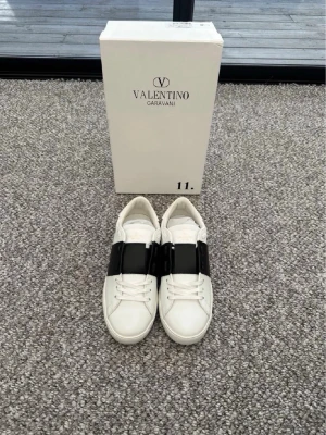 Valentino skor - Snygga vita sneakers från Valentino Garavani med bred mörkblå rem över vristen. Skorna är i läder och har en klassisk låg siluett med diskreta nitar på sulan. Perfekta för dig som vill ha en stilren och exklusiv look.