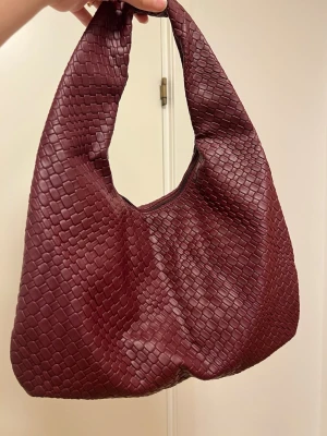 Vinröd handväska - Snygg vinröd handväska med flätad struktur och mjuk, böljande form. Väskan har ett bredare handtag och stängs med dragkedja. Perfekt accessoar för dig som gillar trendiga detaljer och en unik look. Materialet ser ut som konstläder.