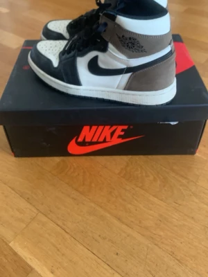 Nike Air Jordan 1 High Mocha sneakers - Snygga Nike Air Jordan 1 High Mocha sneakers med svart, vit och brun färgkombination. Klassisk hög siluett med svart swoosh, svarta skosnören och detaljer i mocka och läder. Perfekta för dig som gillar streetwear och ikoniska sneakers.