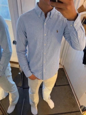 Ljusblå skjorta från Ralph Lauren - Klassisk ljusblå skjorta från Ralph Lauren med broderad logga på bröstet. Skjortan har knappar hela vägen, button-down krage och lång ärm. Perfekt för en stilren och avslappnad look.
