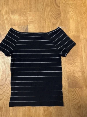 Svart off shoulder topp från Kappahl - Snygg svart off shoulder topp från Kappahl med vita tunna horisontella ränder. Kortärmad och figurnära modell, perfekt till jeans eller kjol. Mjuk och stretchig bomullsblandning som sitter skönt hela dagen.