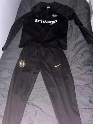 Chelsea svart träningsset Nike - Snyggt svart träningsset från Nike med Chelsea FC-logga och Trivago-tryck på tröjan. Setet består av långärmad tröja och matchande byxor, båda med guldiga detaljer och Nike swoosh. Perfekt för fotbollsträning eller chill. Byten fungerar. FRAKTAR EJ!!! möta bara!