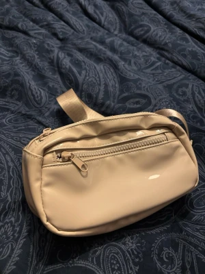 Beige magväska med justerbart band - Snygg beige magväska i glansigt material med justerbart band och dragkedja framtill. Perfekt för att bära det viktigaste och enkel att matcha med olika outfits. Praktisk och stilren design.