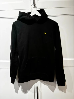 Svart hoodie från Lyle & Scott - Svart hoodie från Lyle & Scott med klassisk gul logga broderad på bröstet. Hoodien har en stor huva och en magficka framtill. Tillverkad i mjukt bomullsmaterial som är skönt att ha på sig. Perfekt för en avslappnad och stilren look. Nypris (750)