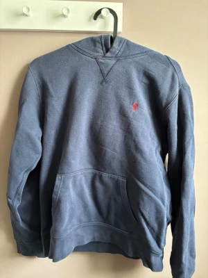 Blå hoodie från Polo Ralph Lauren - Mörkblå hoodie från Polo Ralph Lauren med klassisk röd broderad logga på bröstet. Hoodien har känguruficka framtill, ribbade muddar och en skön huva. Perfekt för en avslappnad och stilren look. Storlek 14-16 men passar XS. Hör av dig vid funderingar om pris eller annat!