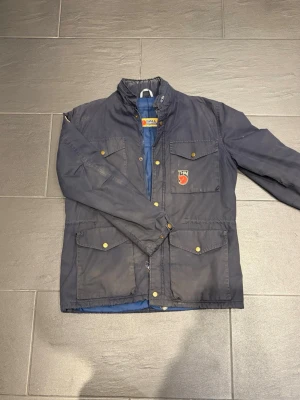 Vintage jacka från Fjällräven - Klassisk mörkblå jacka från Fjällräven stl M, med fyra stora fickor framtill, tryckknappar i guld och broderade logotyper på bröstet och ärmen. Jackan har hög krage och justerbar nederkant med dragsko. Perfekt för uteliv och vardag. Lite smutsig Defekter längst ner på ärmsömen. Nypris 4500kr, säljer för 2000kr, pris kan diskuteras. 