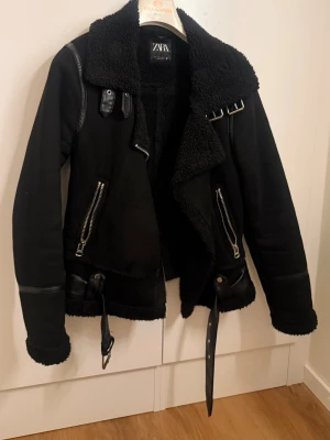Svart skinnjacka med fuskpäls från Zara - Svart skinnjacka från Zara med fuskpälsfoder och stora slag. Jackan har dragkedjor, bälte i midjan och detaljer i skinnimitation. Två snedställda fickor med dragkedja och coola spännen vid kragen ger en edgy vibe.