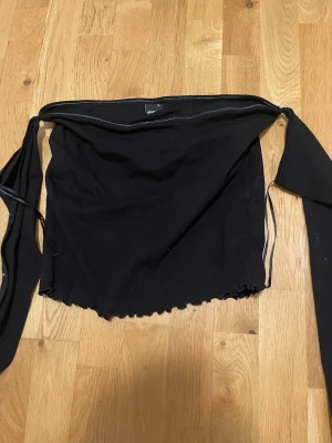 Ginatricot Svart omlottopp med långa band - Trendig svart omlottopp med långa knytband som kan stylas på flera sätt. Toppen är axelbandslös och har en lätt croppad passform med vågig nederkant. Perfekt för att skapa en cool och personlig look.