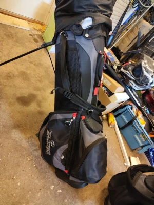 Svart och grå golfbag med stativ - Golfbag i svart och grått med röda detaljer från Spalding. Väskan har flera dragkedjefack, bärrem och inbyggda stativben. Tillverkad i slitstark syntet och har ett skyddande överdrag för klubborna. Två hål som ni ser i bilderna men inget som påverkar användningen