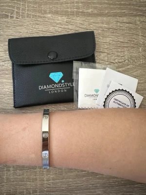 Silverfärgat armband med stenar från Diamond Style - Snyggt och stilrent armband i silverfärg från Diamond Style London. Armbandet är stelt och har infällda glittrande stenar runt om. Kommer med originalförpackning, putsduk och äkthetsintyg. Perfekt accessoar för att lyfta din outfit. OBS: FRAKTAR INOM 24H!🩷 