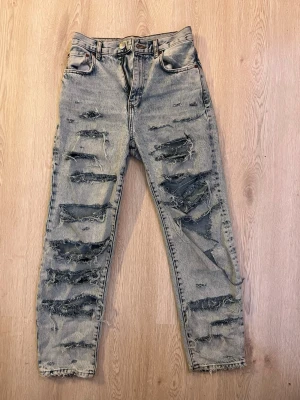 Grå slitna mom jeans med hål - Säljer ett par grå mom jeans med hög midja och massor av slitningar och stora hål framtill. Jeansen har klassisk femficksmodell och raka ben. Perfekta för en edgy och trendig streetstyle-look.