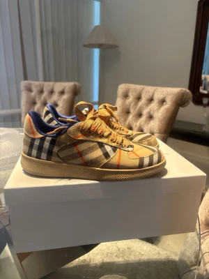 Burberry rutiga sneakers låga - Säljer ett par klassiska Burberry sneakers med det ikoniska beige, svarta och röda rutmönstret. Skorna har lågt skaft, beige sula och matchande snören. Insidan har blå detaljer och ovandelen är i textil och läder. Perfekta för dig som gillar lyxig streetstyle.