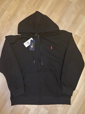 Svart hoodie från Polo Ralph Lauren - Svart hoodie med dragkedja från Polo Ralph Lauren. Klassisk design med huva, känguruficka och röd broderad logga på bröstet. Mjukt material och ribbade muddar vid ärmslut och nederkant. Perfekt för en avslappnad och stilren look.