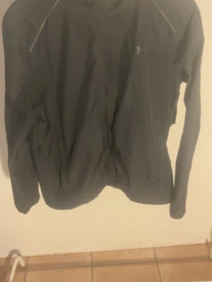 Svart vindjacka från Under Armour - Snygg svart vindjacka från Under Armour med huva och reflekterande detaljer. Jackan har dragkedja framtill och logga på bröstet samt stor text på ryggen. Perfekt för blåsiga dagar och sportiga outfits.