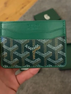 Grön kortplånbok - Snygg grön kortplånbok från med klassiskt mönster och flera kortfack. Plånboken har en unik design med signaturtryck i vitt och gult på framsidan. Perfekt för att hålla ordning på dina kort.
