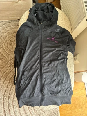 Svart hoodie med dragkedja - Säljer en svart hoodie med dragkedja och huva från. Hoodien har fickor framtill och tryck i lila och vitt på bröstet. Perfekt för gymmet eller chill dagar. Materialet känns mjukt och stretchigt, och modellen är klassisk med ribbade muddar.