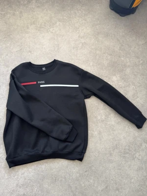 Svart sweatshirt med Paris-tryck - Stilren svart sweatshirt med rund hals och med texten 'PARIS' på bröstet. storlek L men passar även som M. Tröjan har långa ärmar och ribbade muddar vid ärmslut och nederkant. Perfekt för en avslappnad och trendig look. Använd 1-2 ggr. 