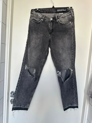 Grå slitna girlfriend jeans H&M - Snygga gråa girlfriend jeans från H&M med låg midja och ankellängd. Jeansen har slitna detaljer och hål på båda knäna samt råa benslut. Tillverkade i denim med återvunna fibrer för en cool och hållbar look.