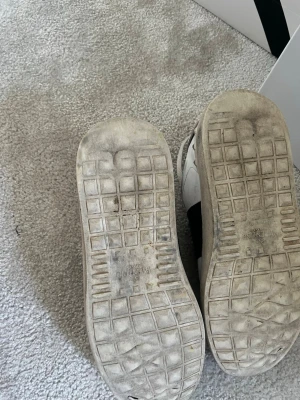 Valentino Garavani vita sneakers - Snygga vita sneakers från Valentino Garavani med en bred svart rem över sidan. Klassisk låg modell med rund tå och vita skosnören. Yttermaterial i läder och gummisula med rutmönster. Perfekta för en trendig streetwear-look.