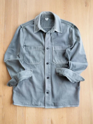Grå/Grön Overshirt från Arket - Stilren grå/grön overshirt i kraftigt bomullstyg med två stora bröstfickor och svarta knappar framtill. Klassisk krage och raka ärmar som kan vikas upp för en avslappnad look. Perfekt lager-på-lager-plagg för en trendig streetstyle.