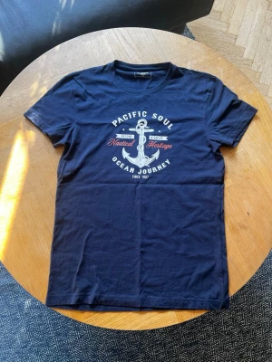 Marinblå t-shirt från Dressmann  - Säljer en marinblå t-shirt från Dressmann med tryck framtill som har ankare och texten 'Pacific Soul Ocean Journey'. T-shirten har rund hals och korta ärmar. Perfekt för dig som gillar lite mer laidback stil.