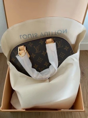 Louis Vuitton Monogram handväska - Snygg handväska från Louis Vuitton i klassiskt brunt monogrammönster med guldiga metalldetaljer och ljusa läderhandtag. Väskan har rundad form och levereras med axelrem och tillbehör. Perfekt för dig som gillar lyxig och tidlös design.