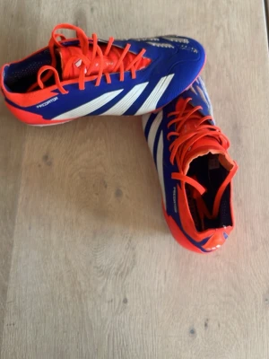 Adidas Predator blå fotbollsskor - Säljer ett par Adidas Predator elite fotbollsskor i blått med vita detaljer och röda snören samt hälparti. Skorna har klassisk låg profil och är tillverkade i syntetmaterial för bra grepp och kontroll på planen. Perfekta för dig som vill sticka ut på matchen.