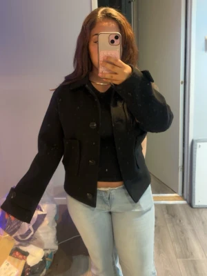 Svart kort jacka med stora knappar - Snygg svart jacka med kortare passform och stora knappar framtill. Jackan har två stora fickor och klassisk krage. Perfekt till jeans eller kjol för en clean look. Materialet känns tjockt och värmande, och ärmarna är långa med knappdetalj vid mudden.