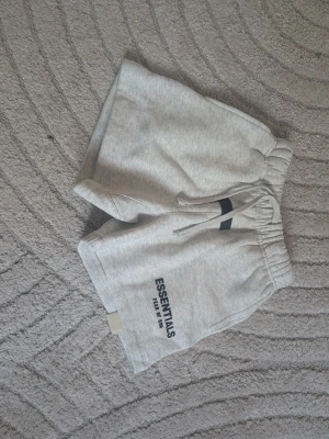 Gråa Essentials shorts med tryck - Snygga ljusgrå shorts från Essentials med svart tryck på ena benet. Mjuk bomullsblandning, elastisk midja med dragsko och avslappnad passform. Perfekta för chill dagar eller streetwear-stil.