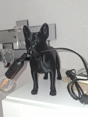 Svart bulldog lampa - Säljer en unik bordslampa formad som en bulldog i matt svart. Lampan har en glödlampa som hunden håller i munnen och en svart sladd med stickkontakt. Perfekt som cool inredningsdetalj för dig som gillar hundar och modern design.
