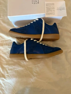 Blå sneakers Maison Margiela - Snygga blå sneakers från Maison Margiela med ovandel i mocka och läder, vita skosnören och klassisk gummisula i brunt. Låg modell med stilren design och diskreta detaljer. Perfekta för dig som vill ha något unikt och trendigt i garderoben.