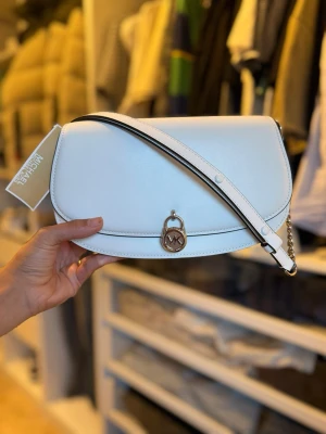 Vit axelremsväska Michael Kors - Snygg vit handväska från Michael Kors i slätt läder med silverfärgad MK-detalj framtill. Väskan har en justerbar axelrem med kedjedetalj och stilren, halvmåneformad design. Perfekt för att lyfta din outfit med en clean och modern look.