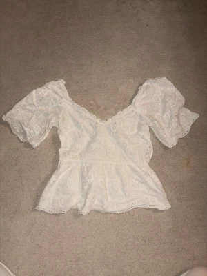 Vit blus med spets och puffärm - Söt vit blus med v-ringning, puffiga korta ärmar och fina spetsdetaljer. Blusen har broderat mönster och volangkant nedtill för en romantisk vibe. Perfekt till jeans eller kjol för en trendig look.