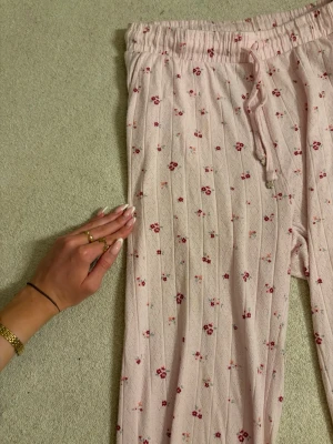 Rosa pyjamasbyxor med blommönster - Söta pyjamasbyxor i ljusrosa med små röda blommor och diskreta ränder. Byxorna har resår i midjan med dragsko och raka ben med vågig kant längst ner. Perfekta för mysiga kvällar hemma.