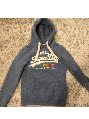 SuperDry Rare Hoodie - Säljer en blå hoodie från Superdry med vit logga och japansk text på bröstet. Hoodien har stora fickor framtill, ribbade muddar och en rejäl huva med vita snören. Perfekt för en avslappnad och cool stil.