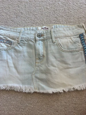 Ljus jeanskjol från Hollister - Kort ljusblå jeanskjol från Hollister med fransad nederkant och broderade detaljer i blått och orange vid fickorna. Klassisk femficksmodell med bälteshällor och knappstängning framtill. Perfekt för en avslappnad och trendig look.💓