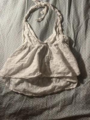Vit broderad halterneck topp - Säljer en söt vit halterneck topp med broderade detaljer och knyt i nacken. Toppen har v-ringning och är lätt utsvängd nedtill, vilket ger en luftig och somrig känsla. Perfekt för varma dagar och enkel att styla med jeans eller kjol.