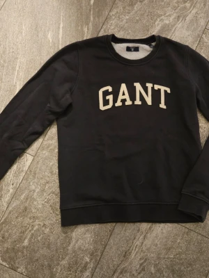 Blå sweatshirt från GANT - Klassisk blå sweatshirt från GANT med rund halsringning och stora broderade bokstäver i vitt framtill. Tröjan har långa ärmar och ribbade muddar vid ärmslut och nederkant. Perfekt för en avslappnad och stilren look.