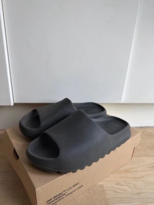 Svarta Adidas tofflor slides - Chunky svarta slides från Adidas med den klassiska trestrecksloggan i sulan. Tofflorna har en clean, minimalistisk look och är tillverkade i syntetmaterial med en tjock, räfflad sula för extra komfort.