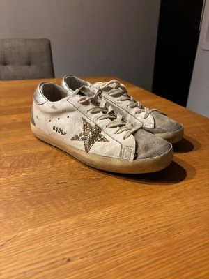 Golden Goose skor - Golden Goose sneakers i vitt skinn med grå mockadetaljer och silverfärgad häl. Sidan pryds av en glittrig guldstjärna. Klassisk snörning och platt sula. Perfekta för dig som vill ha en cool och unik look. Pris kan diskuteras 
