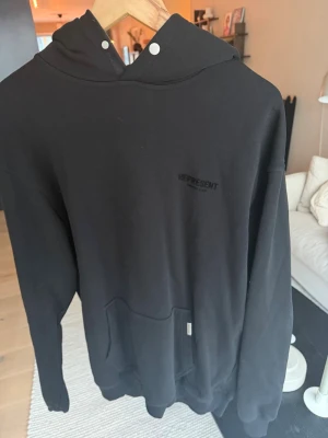 Svart hoodie från Represent - Säljer en svart hoodie från Represent med diskret logga på bröstet. Hoodien har huva med tryckknappar och en stor magficka framtill. Tillverkad i mjukt material och har en avslappnad passform, perfekt för en casual look.