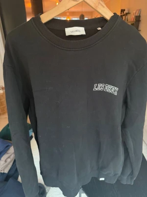 Svart sweatshirt från Les Deux - Svart sweatshirt från Les Deux med broderad vit logga på bröstet. Tröjan har rund halsringning och ribbade muddar vid ärmslut och nederkant. Perfekt för en avslappnad och stilren look.