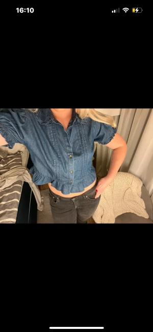 Blå croppad blus med volangkant - Säljer en blå croppad blus i denim-look med korta ärmar, volangkant nedtill och knappar framtill. Blusen har en klassisk krage och lätt puffade ärmar som ger en trendig vibe. Perfekt att styla med höga jeans eller kjol.