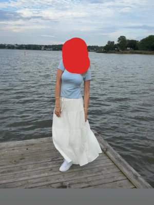 Vit långkjol med knyt i midjan - Säljer en flowy vit långkjol med knytband i midjan. Kjolen har volangdetaljer och är gjord i ett lätt bomullsliknande material som passar perfekt till sommaren. Enkel att styla med sneakers eller sandaler för en chill look.